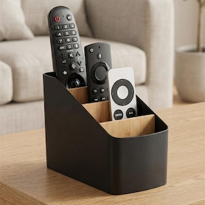 Peut inclure: Un organiseur de télécommandes noir avec trois compartiments, contenant trois télécommandes noires et blanches. L'organisateur est sur une table en bois, avec un canapé et une plante en arrière-plan.
