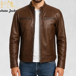 Könnte beinhalten: Braune Lederjacke mit Stehkragen und Reißverschluss. Die Jacke hat zwei Brusttaschen mit Reißverschluss und zwei Seitentaschen. Die Ärmel sind lang und tailliert. Die Jacke wird über einem weißen T-Shirt und einer blauen Jeans getragen. Der Markenname "Stitchure Jack" ist sichtbar.