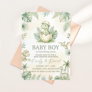 Può includere: Invito per baby shower con un design ad acquerello di un cucciolo di dinosauro che esce da un uovo. L'invito include il testo "Baby Boy is hatching soon" e i dettagli dell'evento, compresi i nomi "Emily & David". L'invito è circondato da fogliame verde.