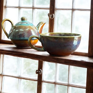 Op de afbeelding: Een geglazuurde keramische theepot met deksel en een grote theekop met oor. De theeset is in tinten bruin, blauw en goud. De theepot en theekop staan op een houten plank voor een raam.