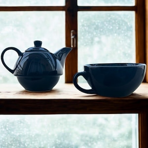 Könnte beinhalten: Eine dunkelblaue Keramik-Teekanne mit passender Tasse und Henkel steht auf einem Holzregal vor einem Fenster. Die Teekanne hat einen runden Körper, einen gebogenen Ausguss und einen kleinen Deckel. Die Tasse hat eine runde Form und einen großen Henkel.