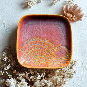 Könnte beinhalten: Quadratischer Keramikteller mit Farbverlauf von Orange nach Rot, mit einem detaillierten orangefarbenen und silbernen Design. Der Teller ist von weißen Blumen und einer pfirsichfarbenen Blume umgeben.