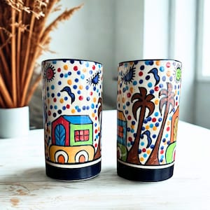 May include: Two cylindrical ceramic glasses with colorful hand-painted designs. The glasses feature a blue rim and base, with a pattern of dots, houses, palm trees, and other shapes in red, blue, green, and yellow. The glasses are on a light wood surface.