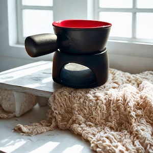Puede incluir: Una olla de fondue negra con interior rojo y asa. La olla se asienta sobre una base negra con una abertura. La olla está sobre una superficie blanca con un tejido de punto beige frente a una ventana.
