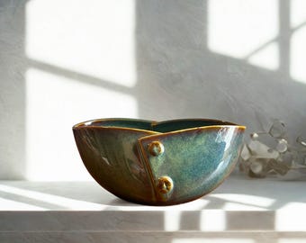 May include: A handcrafted ceramic bowl with a unique design. The bowl features a glossy, teal and brown glaze with two decorative button-like accents. The bowl is placed on a white surface, with sunlight casting shadows in the background.