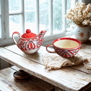 Op de afbeelding: Een rood en wit bloemmotief keramische theepot en kop en schotel set. De theepot heeft een deksel en een handvat, en de kop en schotel heeft een handvat. De set staat op een houten tafel met een neutraal gekleurde doek.