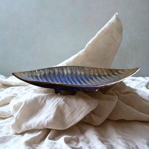 Puede incluir: Un plato de cerámica con forma de hoja con un acabado metálico azul y dorado. El plato tiene una superficie texturizada y está sobre una tela blanca. El plato mide aproximadamente 30 cm de largo.