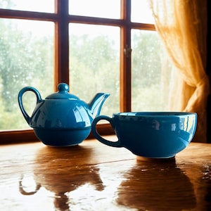 May include: A blue ceramic teapot and teacup on a wooden surface near a window. The teapot has a rounded body, a curved handle, and a spout. The teacup has a large handle. The objects reflect in the shiny wooden surface.