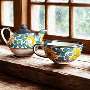 May include: A ceramic teapot and teacup set with a floral design. The teapot has a blue lid and handle, and the cup has a matching blue rim and handle. Both pieces are decorated with yellow flowers and green leaves. The set is placed on a wooden surface.
