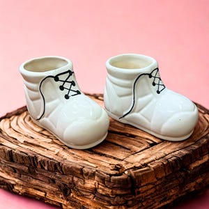 May include: Two white ceramic baby boot planters with black lace detailing. The planters are on a rustic wooden base against a pink background. The boot planters are a charming decorative item.