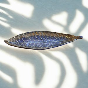 Puede incluir: Un plato decorativo en forma de hoja con acabado metálico. El plato es de color azul oscuro con vetas plateadas y se encuentra sobre una superficie blanca. El plato en forma de hoja es un artículo decorativo.