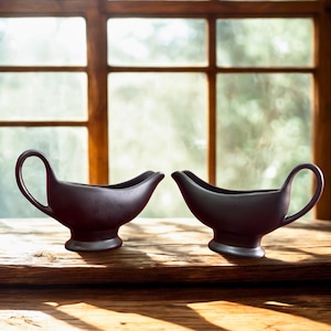 Könnte beinhalten: Zwei dunkelgraue Keramik-Saucierschalen mit Griffen und Ausgüssen stehen auf einer Holzoberfläche. Die Schalen stehen vor einem Fenster mit natürlichem Licht und einem verschwommenen Hintergrund.