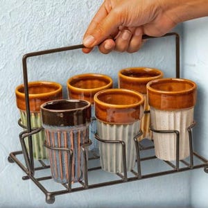 Puede incluir: Un juego de seis vasos de cerámica en un soporte de metal. Los vasos tienen un diseño acanalado con tonos marrones y crema. El soporte de metal es de color marrón oscuro y está sostenido por una mano.