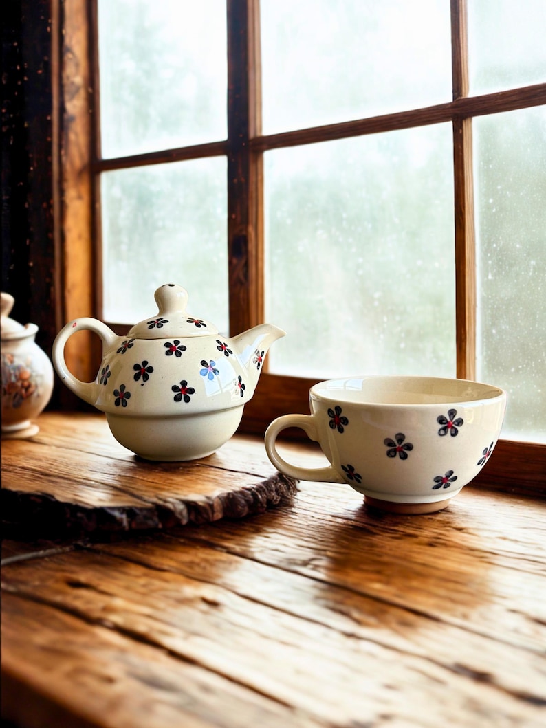 May include: Cream-colored teapot and teacup set with a floral pattern. The teapot has a rounded body, a curved spout, and a small handle. The teacup has a large handle. Both are decorated with small, dark blue and red flowers.