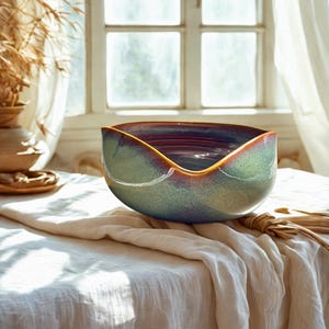 May include: A handcrafted ceramic bowl with a unique, asymmetrical shape. The bowl features a blend of green, blue, and purple hues with a golden rim. It sits on a white linen tablecloth, with natural light illuminating the piece, suggesting a decorative or serving purpose.