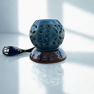 Pode incluir: Um difusor de óleos essenciais de cerâmica azul-petróleo com um topo esférico perfurado e uma base marrom. O difusor elétrico tem um cabo preto e foi projetado para dispersar óleos essenciais.