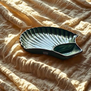 Puede incluir: Un plato verde de cerámica con forma de concha marina y un pequeño compartimento adjunto. El plato tiene una textura acanalada y un acabado brillante. El fondo es una tela texturizada de color amarillo claro.