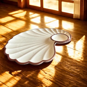 Puede incluir: Una fuente blanca en forma de concha, con un borde marrón y un pequeño plato circular adjunto. La fuente tiene una superficie texturizada con motas sutiles. El diseño recuerda a una concha de vieira. La fuente está sobre una superficie de madera.