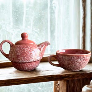 May include: A red speckled ceramic teapot with a matching lid and a handle, next to a matching teacup. The teapot has a spout and sits on a wooden surface. The teacup has a handle and a rounded shape. The background is a window.