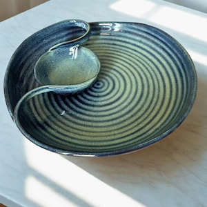 Könnte beinhalten: Eine runde Keramik-Servierschale mit einer tiefblauen und lindgrünen Glasur und einer markanten Spiralstruktur. Die Schale verfügt über einen integrierten, kleineren kreisförmigen Schalenbereich an der Seite, der ein durchgehendes, geschwungenes Randdesign bildet.