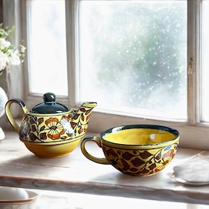 May include: A yellow ceramic teapot and teacup set with a floral design in brown and orange. The teapot has a dark green lid and handle. The teacup has a matching floral pattern and a yellow interior. The set is placed on a wooden surface near a window.