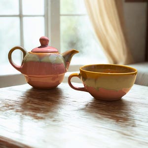 以下が含まれることがあります： 層状の釉薬デザインが施された陶器のティーポットとティーカップ。ティーポットは丸みを帯びた本体、湾曲した注ぎ口、つまみ付きの蓋が特徴です。ティーカップには広い縁とサイドハンドルが付いています。どちらの作品もピンク、ベージュ、黄色の水平な帯模様が特徴です。