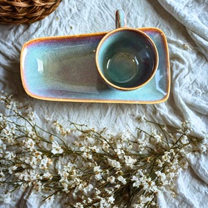 Può includere: Un set di tazza e piattino in ceramica con un vassoio rettangolare. Il set presenta una glassa turchese e iridescente con un bordo marrone. La tazza ha un manico. Il set è su un panno bianco con fiori bianchi e un cestino di vimini.