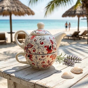 May include: A ceramic tea-for-one set consisting of a teapot stacked on a matching teacup. Both pieces feature a cream-colored base with a repeating pattern of red flowers, black leaves, and thin black swirling vines. A sprig of lavender and two seashells sit nearby.