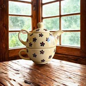 May include: A cream-colored ceramic teapot and teacup set with a floral pattern of red, blue, and black flowers. The set is placed on a wooden table near a window.