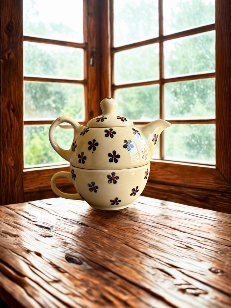 May include: A cream-colored ceramic teapot and teacup set with a floral pattern of red, blue, and black flowers. The set is placed on a wooden table near a window.