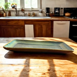 Puede incluir: Una fuente rectangular de cerámica con superficie acanalada. La fuente tiene un esmalte verde claro y azul y está sobre una mesa de madera. El fondo de la cocina incluye una ventana, armarios y electrodomésticos.