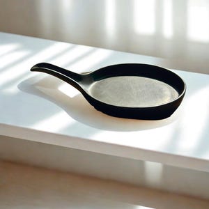Op de afbeelding: Een zwarte serveerschaal met een handvat, rustend op een wit oppervlak. De schaal heeft een afgeronde vorm met een licht verhoogde rand. Het handvat steekt uit de zijkant, wat een strak, modern ontwerp creëert. De schaal is gemaakt van mat materiaal.