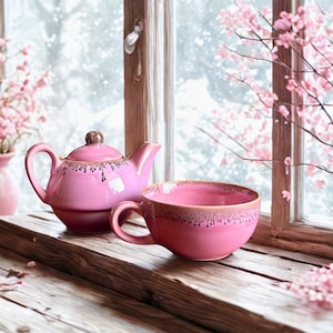 Peut inclure: Un service à thé rose en céramique, composé d'une théière et d'une tasse, avec un bord marron. La théière a un couvercle et une anse, et la tasse a une anse. L'arrière-plan comprend une fenêtre et des branches fleuries roses.