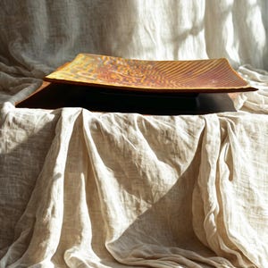 May include: A rectangular, amber ceramic serving platter with a textured surface. The platter sits on a dark, rectangular base, displayed on a cream linen cloth. The design features concentric lines and decorative elements.
