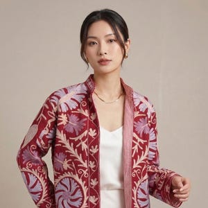 May include: A burgundy jacket with detailed floral embroidery in lavender and cream. The jacket has a mandarin collar and a striped lining. The sleeves are long, and the jacket is open at the front. The jacket is worn over a white dress.