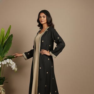 Może przedstawiać: Kobieta w czarnym płaszczu z długim rękawem na beżowej sukience. Płaszcz ma złote haftowane detale i wzory w kształcie gwiazd. Sukienka ma dekolt w serek i zwiewną sylwetkę, odpowiednią na formalne okazje.