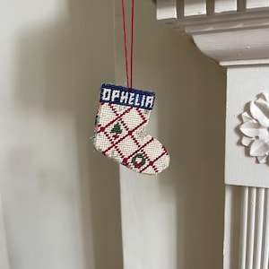 May include: A miniature, hand-stitched Christmas stocking decoration. The stocking features a white base with a red and green diamond pattern. The top band is blue with the name "OPHELIA". A red string is attached for hanging.