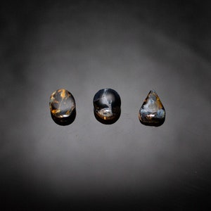 May include: Three polished stones with a blue and gold tiger's eye pattern. The stones are oval, round, and teardrop shaped.
