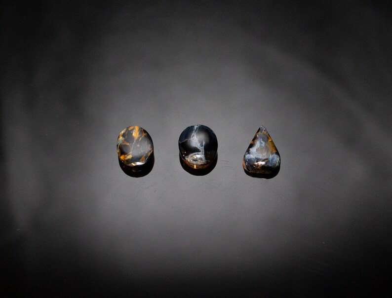 May include: Three polished stones with a blue and gold tiger's eye pattern. The stones are oval, round, and teardrop shaped.