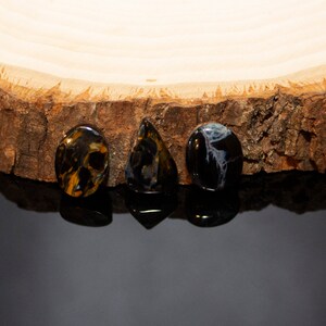 May include: Three gemstone cabochons, one oval, one teardrop, and one round, in shades of blue, brown, and gold, on a wooden surface.
