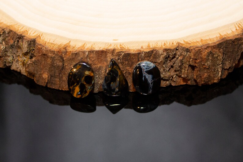 May include: Three gemstone cabochons, one oval, one teardrop, and one round, in shades of blue, brown, and gold, on a wooden surface.