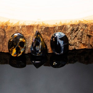 May include: Three gemstone cabochons, one oval, one teardrop, and one round, in shades of blue, black, and gold, on a wooden surface.