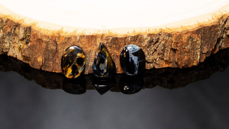 May include: Three gemstone cabochons, one oval, one teardrop, and one round, in shades of blue, black, and gold, on a wooden surface.