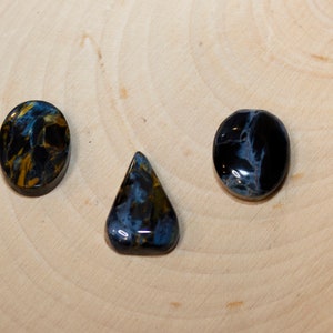 May include: Three blue tiger's eye cabochons in different shapes: oval, teardrop, and round. The stones have a dark blue base with gold and white streaks.