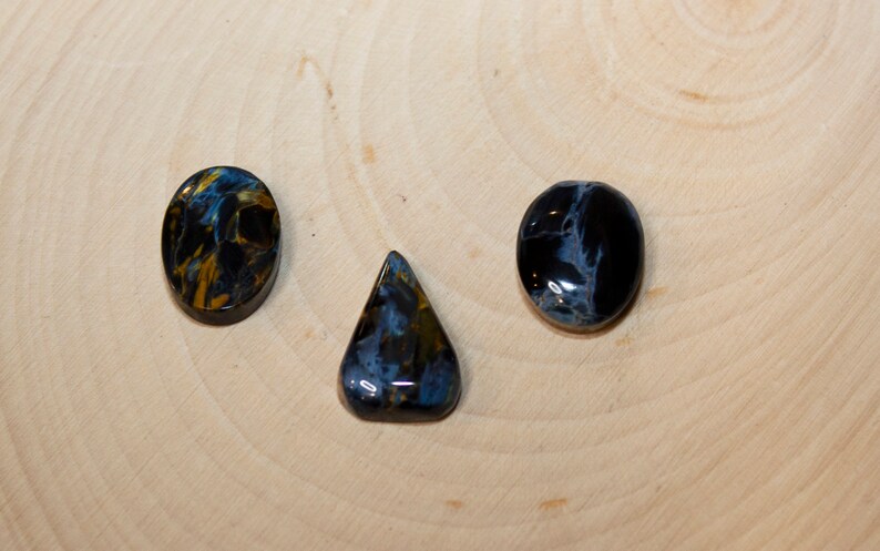 May include: Three blue tiger's eye cabochons in different shapes: oval, teardrop, and round. The stones have a dark blue base with gold and white streaks.