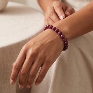 May include: A beaded bracelet featuring round, faceted, deep magenta-colored stones. The beads are uniform in size and strung together in a continuous circular loop, displaying a polished, reflective surface texture throughout the entire piece.
