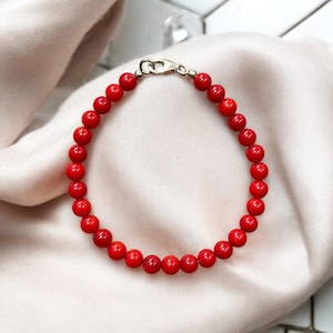 May include: A red beaded bracelet with a silver clasp. The bracelet is made of small, round, vibrant red beads. The bracelet is displayed on a cream-colored, silky fabric.