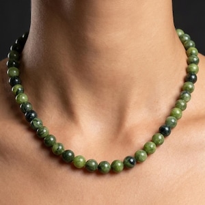 Könnte beinhalten: Eine Halskette mit runden, grünen Perlen mit dunkelgrüner und schwarzer Marmorierung. Die Halskette wird vor einem neutralen Hintergrund präsentiert, der die natürlichen Farbvariationen der Steinperlen hervorhebt. Ein klassisches Schmuckstück.