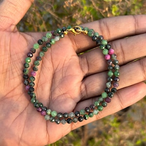 Può includere: Un braccialetto a doppia fila di perline con piccole perline sfaccettate nei toni del verde, nero e rosa. Il braccialetto è assicurato con una chiusura color oro. Il braccialetto è esposto su una mano.