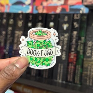 Op de afbeelding: Een sticker met een cartoon pot gevuld met groene bankbiljetten. De pot heeft een lichtbruin deksel en een banner met de tekst "BOOK FUND" in zwarte letters. De sticker heeft een witte rand en steraccenten.